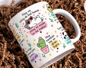 Puede incluir: Una taza de cerámica blanca con ilustraciones coloridas de dibujos animados de un gato con gafas de corazón, un cactus y un aguacate, acompañadas de textos en español como Hoy no estoy de mal humor y estoy en modo no me hables.
