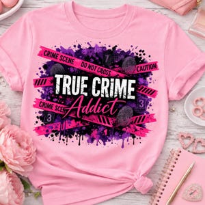 May include: A light pink t-shirt featuring a graphic design with purple and black paint splatters, pink caution tape strips, fingerprint icons, and the text "TRUE CRIME Addict" in white and pink lettering across the center of the chest.