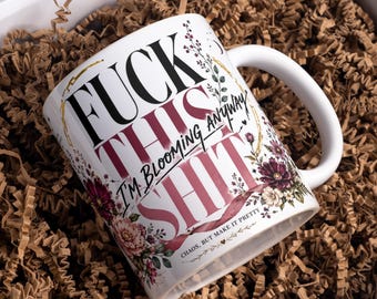 Puede incluir: Una taza de cerámica blanca con un diseño floral de flores rosas, rojas y moradas oscuras. El texto dice FUCK THIS SHIT, I'M BLOOMING ANYWAY y CHAOS, BUT MAKE IT PRETTY en una mezcla de fuentes serif y script en colores negro y rosa.
