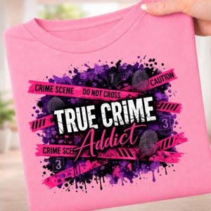 May include: A bright pink t-shirt featuring a graphic design with purple paint splatters, black fingerprint icons, and pink caution tape strips. The text reads "CRIME SCENE DO NOT CROSS" and "TRUE CRIME Addict" in bold white and black lettering.