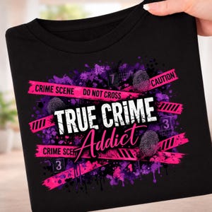 May include: A black t-shirt featuring a graphic design with purple and pink paint splatters, pink crime scene tape with the text "CRIME SCENE DO NOT CROSS", white text reading "TRUE CRIME", pink script text "Addict", and fingerprint icons.