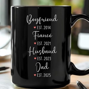 Op de afbeelding: Een zwarte keramische mok met witte tekst die luidt: "Boyfriend EST. 2014, Fiancé EST. 2021, Husband EST. 2023, Dad EST. 2025". Elke titel wordt gevolgd door een rood hart. De mok heeft een grote handgreep.