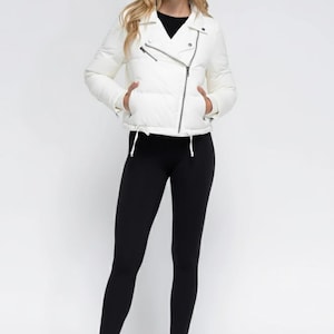 Könnte beinhalten: Eine weiße, gesteppte Jacke mit asymmetrischem Reißverschluss, dazu ein schwarzes Oberteil und schwarze Leggings. Die Jacke hat einen Reverskragen und zwei Vordertaschen. Das Model trägt schwarze Stiefeletten. Der Hintergrund ist weiß.