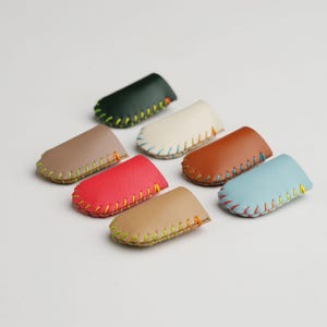 May include: A collection of seven leather key holders in various colors, including green, beige, red, brown, and blue. Each holder is hand-stitched with contrasting thread, showcasing a minimalist design. The key holders are arranged on a white surface.