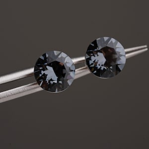 Puede incluir: Dos cristales redondos de color gris oscuro con facetas, sostenidos por unas pinzas de metal plateado. Los cristales presentan un corte brillante con múltiples facetas triangulares que reflejan la luz, creando un aspecto de carbón profundo.