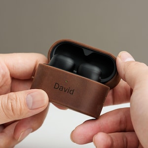 May include: Brown leather earbud case with the name "David" engraved on the front. The case is open, revealing black earbuds inside. The case is held in two hands against a neutral background.