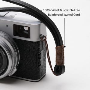 May include: A silver and black camera with a black leather strap. The strap is made of a reinforced waxed cord and is described as 100% silent and scratch-free. The camera lens is visible.