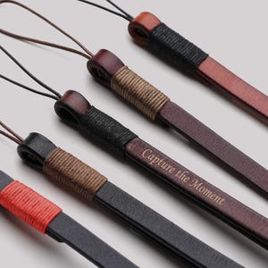 May include: Close-up of several leather camera wrist straps in various colours, including black, brown, and red. Each strap features a wrapped accent and a thin cord for attachment. One strap has the text "Capture the Moment".