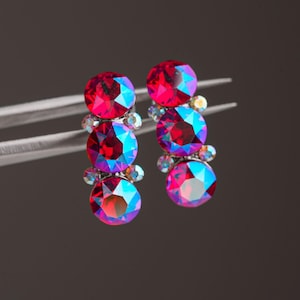 Può includere: Due componenti verticali per orecchini, ciascuno caratterizzato da tre cristalli rotondi sfaccettati di un rosso vibrante con riflessi iridescenti blu e viola. I cristalli sono disposti in fila e fissati in montature di metallo argentato.