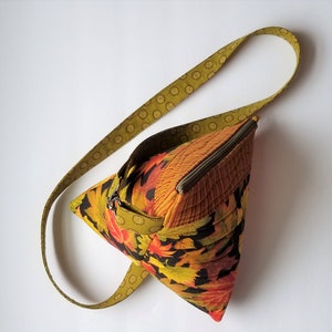 May include: A small, triangular-shaped bag with a zipper closure and a long, patterned strap. The bag is made of a fabric with a fall leaf print in shades of orange, yellow, and brown.
