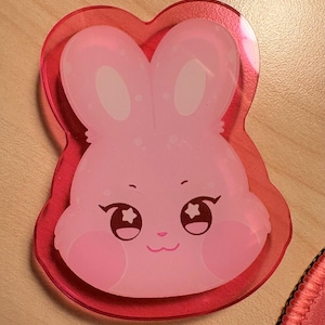 May include: A translucent pink acrylic charm shaped like a rabbit head. The rabbit has large eyes with star-shaped highlights, a small nose, and a smiling mouth. The design is layered over a slightly larger, solid transparent red acrylic backing.