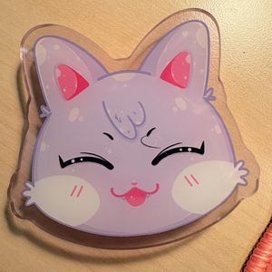 May include: A clear acrylic charm featuring a stylized light purple cat head with large pointed ears, pink inner ears, white cheek patches, and a happy expression with closed eyes, a small pink nose, and a tiny tongue sticking out.