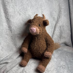 May include: A brown knitted plush toy shaped like a cow with a fuzzy texture, small horns, and a pink nose. The stuffed animal is in a seated position with dark brown limbs and a lighter brown snout, featuring small black eyes and a soft, textured body.