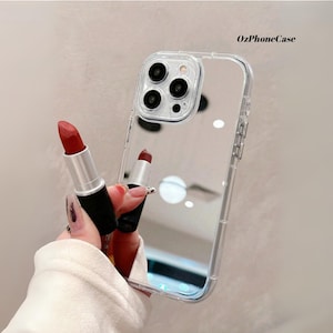 May include: Clear phone case with a mirrored back, featuring a red lipstick. The case has a protective bumper around the camera and edges. The lipstick is held in front of the case, showcasing its color and design. The text "OzPhoneCase" is visible.