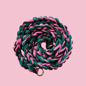 May include: A braided rope leash in a spiral pattern, featuring pink, green, and black fabric. The leash has a gold-colored metal ring at the end. The background is a solid light pink.
