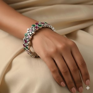 以下が含まれることがあります： フローラルデザインのシルバーカラーのバングルブレスレット。ブレスレットには、緑、赤、透明の宝石が交互に配置されています。ブレスレットは手首に着用されています。