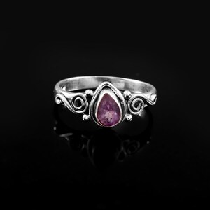 Puede incluir: Un anillo de plata con una piedra preciosa de color púrpura claro en forma de pera. El anillo presenta espirales decorativas en los laterales y una banda lisa. La piedra está engastada en un bisel, sobre fondo negro.