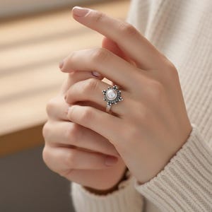 May include: A silver ring featuring a central white pearl surrounded by a detailed, ornate design. The ring's band is thin and delicate, and the overall style is vintage-inspired.