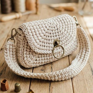 May include: A cream-colored, crocheted handbag with a flap closure and a long strap. The bag features a circular metal clasp and hardware. The bag is sitting on a wooden surface, with sewing notions in the background.