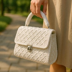 May include: Cream-colored handbag with a woven pattern and a braided handle. The bag features a silver-colored clasp and a flap closure. The bag is held by a person, with a blurred background of greenery.