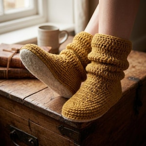 May include: A pair of mustard yellow crochet slipper boots with a textured, slouchy knit design and a light-colored, flat sole. The boots feature a thick, ribbed cuff that extends above the ankle, providing a cozy, handcrafted appearance.