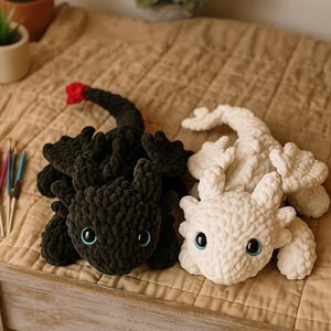 May include: Two handmade crochet dragon toys. One is black with a red tail tip, the other is white. Both have large, round blue eyes. They are lying on a tan quilted surface.