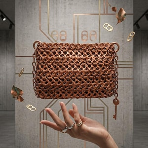 May include: A copper-colored handbag made from recycled pull tabs, with a chain strap and a key charm. The bag is suspended in mid-air, with several pull tabs and a leaf floating around it. The background is a concrete wall with gold geometric lines.