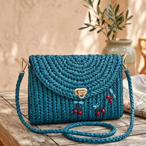 May include: Teal-colored woven handbag with a braided shoulder strap. The bag features a flap closure with a gold-tone clasp and is adorned with cherry blossom accents. The bag is sitting on a wooden surface.