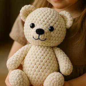 May include: Cream-colored crochet teddy bear with black eyes and a black nose. The bear has a smiling mouth and is being held by a person. The bear's body is made of a textured, looped yarn.