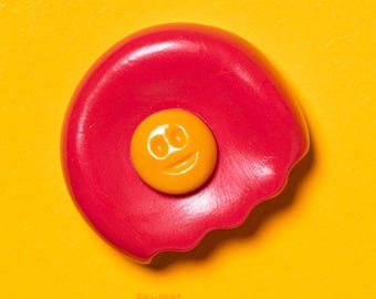 May include: A red, egg-shaped object with a yellow smiley face in the center, set against a yellow and red background. The object has a bite taken out of it. The text "not obliged to smiley" is in the upper right corner.