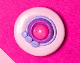 May include: A round, white and pink brooch with concentric circles of purple and pink. Three small, light purple beads are on the left side. The background is a diagonal split of pink and light pink.