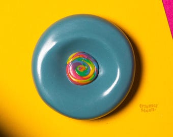 May include: A teal blue, circular object with a rainbow-colored swirl in the center, set against a vibrant yellow and pink background. The text "swirly feelings of (teal) blue" is at the top.