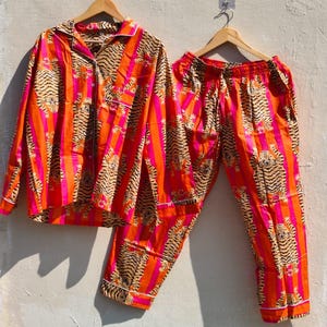 K&ouml;nnte beinhalten: Ein zweiteiliges Pyjama-Set mit einem lebendigen Tigerstreifenmuster. Das Set beinhaltet ein Hemd mit Kn&ouml;pfen und eine passende Hose. Der Pyjama hat ein auff&auml;lliges Design mit orangefarbenen, rosa und schwarzen Streifen und wird auf Holzb&uuml;geln pr&auml;sentiert.