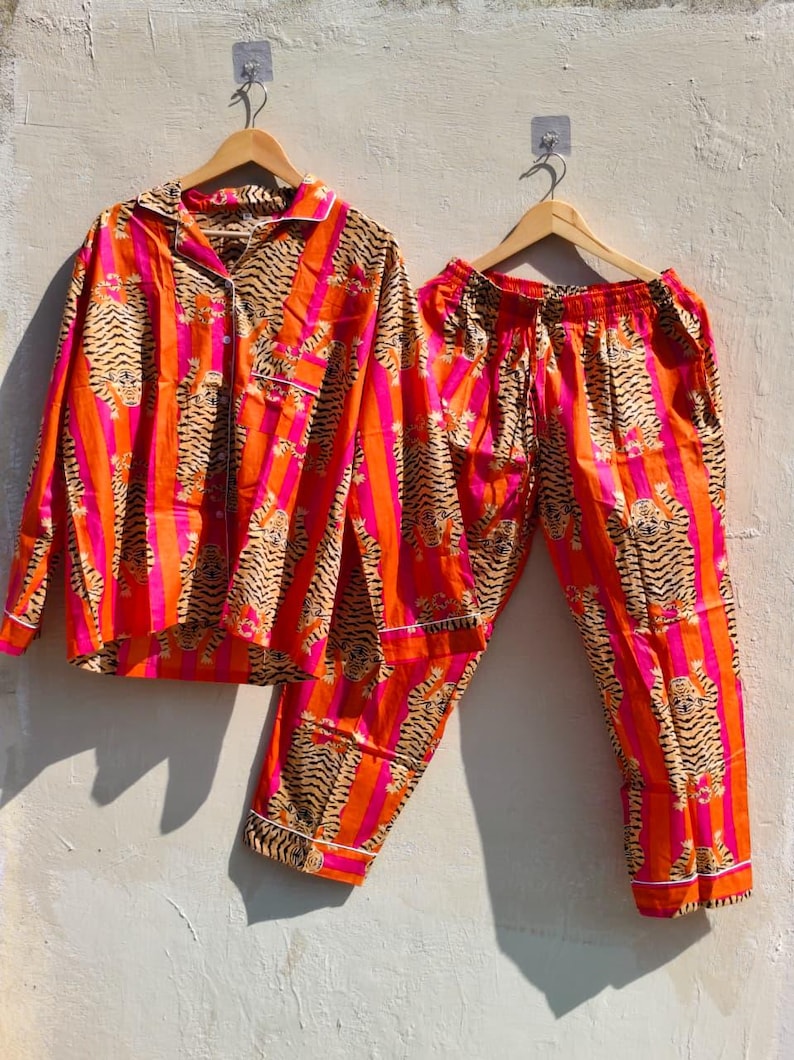 K&ouml;nnte beinhalten: Ein zweiteiliges Pyjama-Set mit einem lebendigen Tigerstreifenmuster. Das Set beinhaltet ein Hemd mit Kn&ouml;pfen und eine passende Hose. Der Pyjama hat ein auff&auml;lliges Design mit orangefarbenen, rosa und schwarzen Streifen und wird auf Holzb&uuml;geln pr&auml;sentiert.