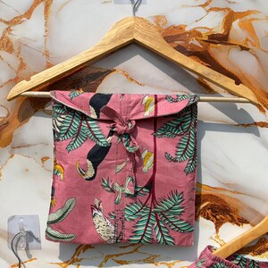 Może przedstawiać: R&oacute;żowy zestaw piżamowy z nadrukiem tukan&oacute;w i palm, zawieszony na drewnianym wieszaku. Zestaw zawiera koszulę zapinaną na guziki i szorty. Tkanina ma powtarzający się wz&oacute;r tukan&oacute;w, liści i element&oacute;w tropikalnych.