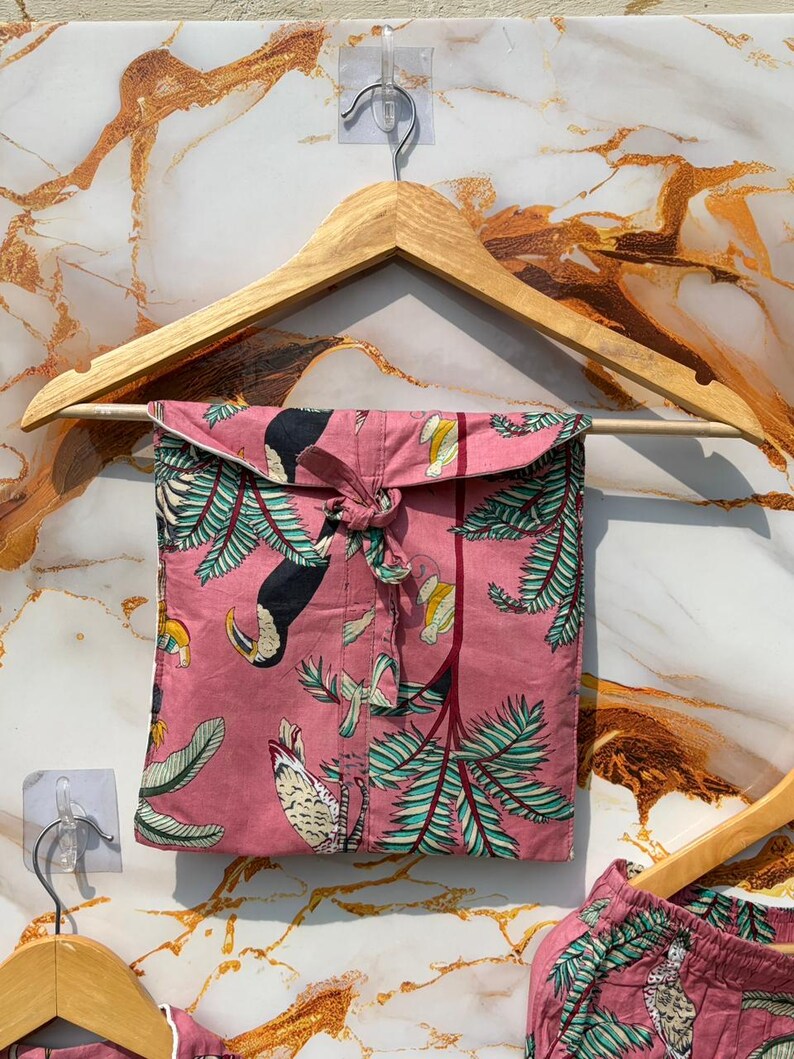 Może przedstawiać: R&oacute;żowy zestaw piżamowy z nadrukiem tukan&oacute;w i palm, zawieszony na drewnianym wieszaku. Zestaw zawiera koszulę zapinaną na guziki i szorty. Tkanina ma powtarzający się wz&oacute;r tukan&oacute;w, liści i element&oacute;w tropikalnych.