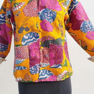 May include: A quilted jacket featuring a vibrant mustard yellow base with a multi-colored floral and abstract pattern in shades of pink, blue, and green. The jacket has a mandarin collar, long sleeves, a front button closure, and two pockets.