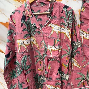 Może przedstawiać: R&oacute;żowy zestaw piżamowy z nadrukiem w dżungli. Zestaw zawiera koszulę zapinaną na guziki i pasujące spodnie. Nadruk przedstawia białe lamparty, zebry, tukany i palmy. Koszula ma białe wykończenia i kieszeń na piersi.