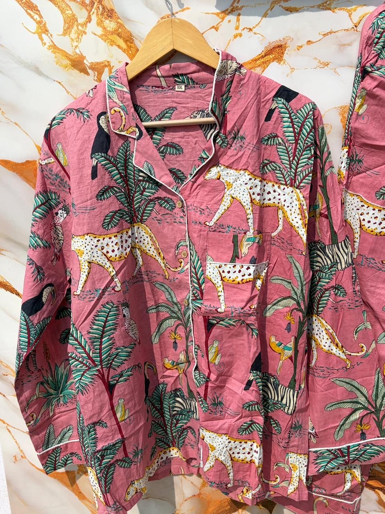 Może przedstawiać: R&oacute;żowy zestaw piżamowy z nadrukiem w dżungli. Zestaw zawiera koszulę zapinaną na guziki i pasujące spodnie. Nadruk przedstawia białe lamparty, zebry, tukany i palmy. Koszula ma białe wykończenia i kieszeń na piersi.