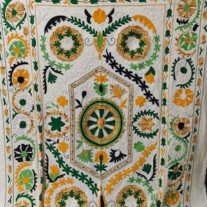 Könnte beinhalten: Weißer Stoff mit aufwendiger Stickerei in Grün, Gelb und Schwarz. Das Design zeigt florale und geometrische Muster, darunter kreisförmige Motive und stilisierte Blätter. Die Stickerei ist detailliert und bedeckt die gesamte Oberfläche.