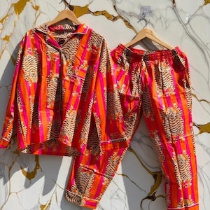 K&ouml;nnte beinhalten: Ein lebendiges Pyjama-Set mit einem Tigerstreifenmuster in Orange, Pink und Braun. Das Set beinhaltet ein Hemd mit Kn&ouml;pfen und eine passende Hose, beide mit wei&szlig;em Besatz. Die Pyjamas werden auf Holzb&uuml;geln pr&auml;sentiert.