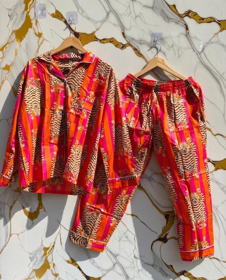 K&ouml;nnte beinhalten: Ein lebendiges Pyjama-Set mit einem Tigerstreifenmuster in Orange, Pink und Braun. Das Set beinhaltet ein Hemd mit Kn&ouml;pfen und eine passende Hose, beide mit wei&szlig;em Besatz. Die Pyjamas werden auf Holzb&uuml;geln pr&auml;sentiert.