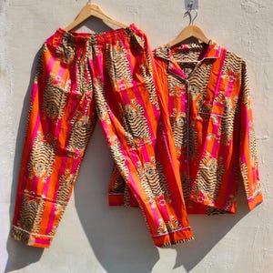 K&ouml;nnte beinhalten: Ein zweiteiliges Pyjama-Set mit einem Tigerstreifenmuster in Orange, Pink und Beige. Das Set beinhaltet ein Hemd mit Kn&ouml;pfen und wei&szlig;em Besatz sowie eine passende Hose. Die Pyjamas h&auml;ngen an Holzb&uuml;geln.