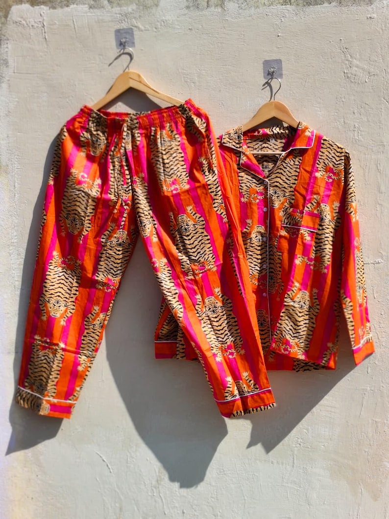 K&ouml;nnte beinhalten: Ein zweiteiliges Pyjama-Set mit einem Tigerstreifenmuster in Orange, Pink und Beige. Das Set beinhaltet ein Hemd mit Kn&ouml;pfen und wei&szlig;em Besatz sowie eine passende Hose. Die Pyjamas h&auml;ngen an Holzb&uuml;geln.