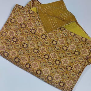 May include: A folded, reversible quilt with a floral pattern. The top layer has a mustard yellow background with a repeating pattern of maroon and cream-coloured floral medallions. The reverse side is a solid mustard yellow with white stitching.