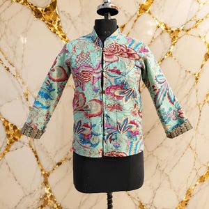 May include: A light blue quilted jacket featuring a vibrant floral pattern in shades of pink, red, and dark blue. The jacket has a mandarin collar, long sleeves with patterned cuffs, a front button closure, and two front pockets on the lower section.