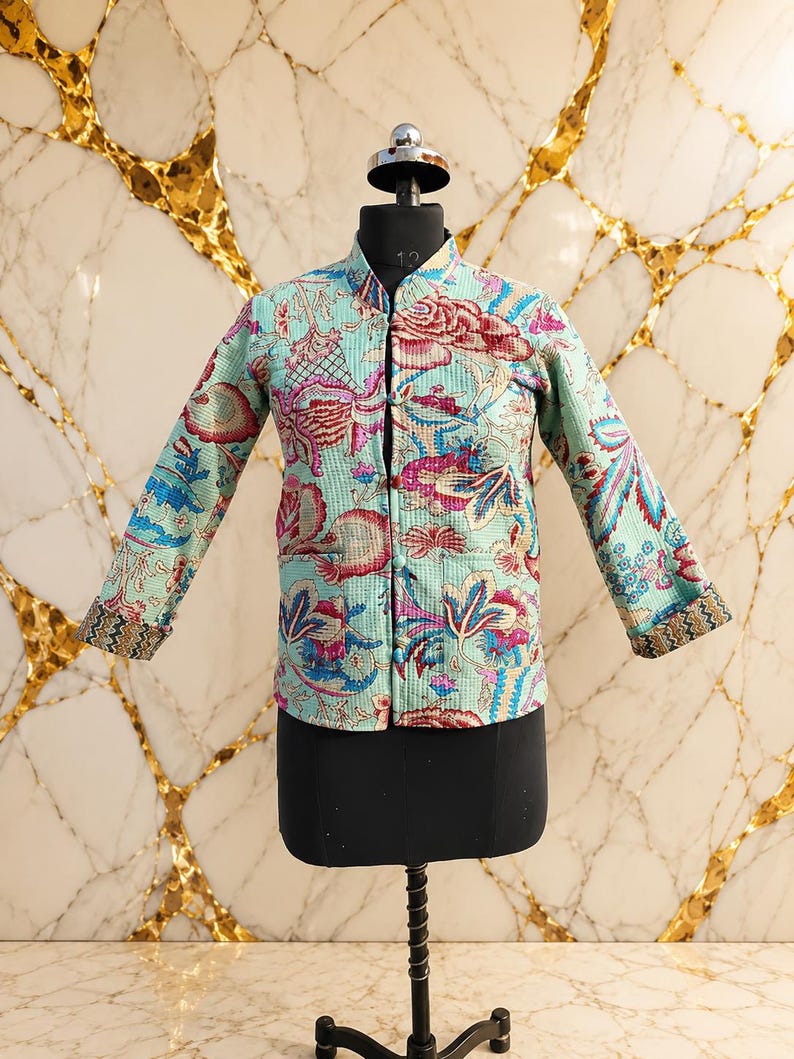 May include: A light blue quilted jacket featuring a vibrant floral pattern in shades of pink, red, and dark blue. The jacket has a mandarin collar, long sleeves with patterned cuffs, a front button closure, and two front pockets on the lower section.