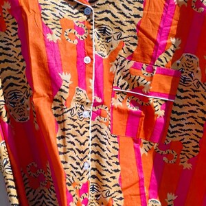 K&ouml;nnte beinhalten: Nahaufnahme eines Pyjama-Oberteils mit Kn&ouml;pfen und einem lebendigen Tigerdruck. Der Stoff weist orange und pinkfarbene vertikale Streifen mit einem sich wiederholenden Muster von Tigern in Braun- und Schwarzt&ouml;nen auf. Wei&szlig;e Paspeln am Kragen, der Tasche und der Knopfleiste.