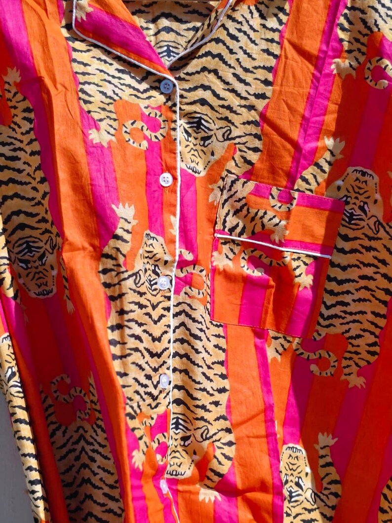 K&ouml;nnte beinhalten: Nahaufnahme eines Pyjama-Oberteils mit Kn&ouml;pfen und einem lebendigen Tigerdruck. Der Stoff weist orange und pinkfarbene vertikale Streifen mit einem sich wiederholenden Muster von Tigern in Braun- und Schwarzt&ouml;nen auf. Wei&szlig;e Paspeln am Kragen, der Tasche und der Knopfleiste.