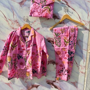 May include: Pink floral pajama set, including a button-down shirt, pants, and a matching pouch. The set features a vibrant floral pattern with green and yellow accents. The shirt and pants are displayed on wooden hangers.